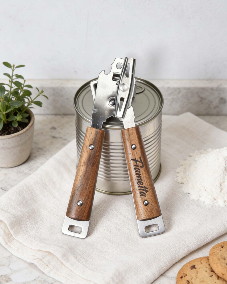 May include: A stainless steel can opener with wooden handles. The handles are engraved with the name "Fiametta". The can opener is positioned over a metal can, with a white cloth, flour, and cookies in the background.
