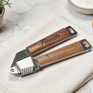 May include: A stainless steel and wood garlic press with the name "Fiametta" engraved on the handle. The press is on a white cloth, with a marble surface in the background. The wooden handles have a natural wood grain.
