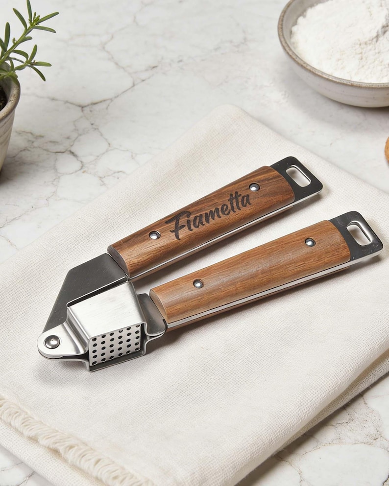 May include: A stainless steel and wood garlic press with the name "Fiametta" engraved on the handle. The press is on a white cloth, with a marble surface in the background. The wooden handles have a natural wood grain.