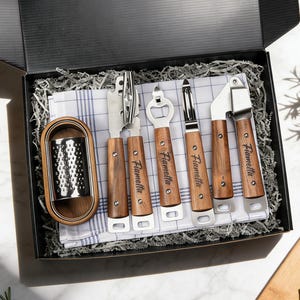 May include: A boxed kitchen utensil set featuring a cheese grater, can opener, bottle opener, peeler, and garlic press. Each tool has a wooden handle and the name "Fiametta" printed on it. The set rests on a blue and white checkered cloth.
