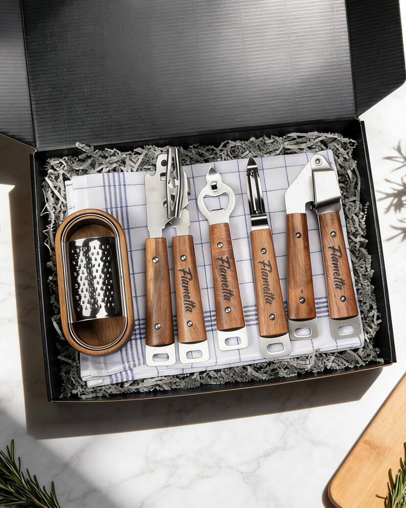 May include: A boxed kitchen utensil set featuring a cheese grater, can opener, bottle opener, peeler, and garlic press. Each tool has a wooden handle and the name "Fiametta" printed on it. The set rests on a blue and white checkered cloth.