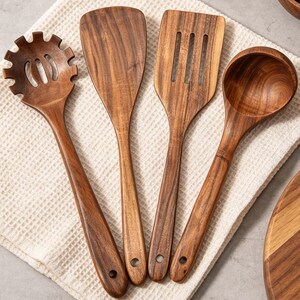 K&ouml;nnte beinhalten: Ein Set aus vier h&ouml;lzernen K&uuml;chenutensilien, darunter ein Schlitzl&ouml;ffel, ein Pfannenwender und eine Sch&ouml;pfkelle, auf einem wei&szlig;en Waffelstoff. Die Utensilien sind aus warmtonigem Holz mit sichtbarer Maserung gefertigt und haben jeweils ein Loch im Griff.