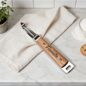 May include: A stainless steel and wood peeler with the name "Fiametta" engraved on the wooden handle. The peeler is lying on a white cloth on a marble countertop, with a small plant and a bowl of flour in the background.