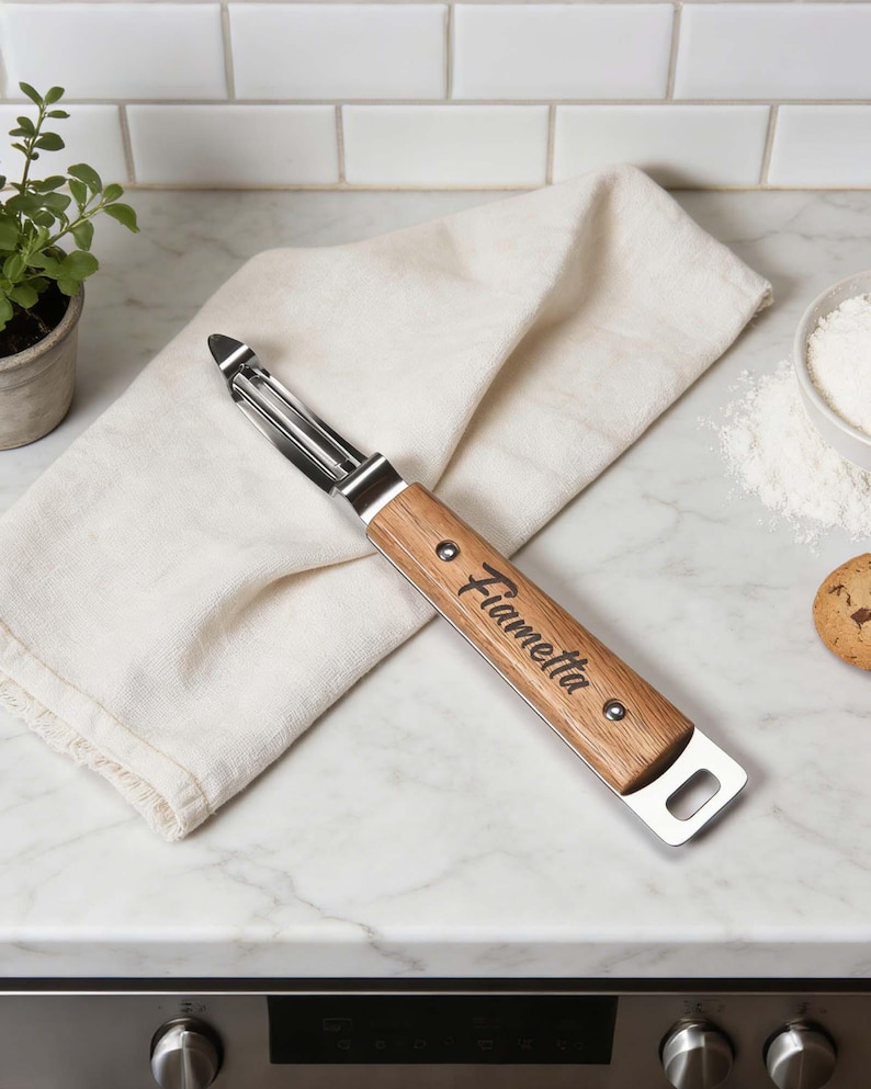 May include: A stainless steel and wood peeler with the name "Fiametta" engraved on the wooden handle. The peeler is lying on a white cloth on a marble countertop, with a small plant and a bowl of flour in the background.