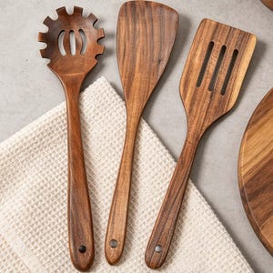 K&ouml;nnte beinhalten: Ein Set aus drei h&ouml;lzernen K&uuml;chenutensilien: ein Nudell&ouml;ffel, ein Pfannenwender und ein Schlitzwender. Die Utensilien bestehen aus dunkelbraunem Holz mit sichtbarer Maserung und haben eine glatte Oberfl&auml;che. Sie liegen auf einem wei&szlig;en Waffel-Tuch.