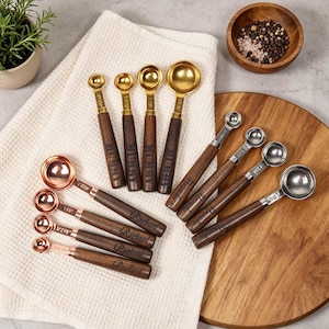 May include: A collection of measuring spoons with wooden handles and metal measuring cups. Some spoons have gold-colored cups, while others are copper-colored. The handles are engraved with names. The spoons are arranged on a white cloth and a wooden cutting board.