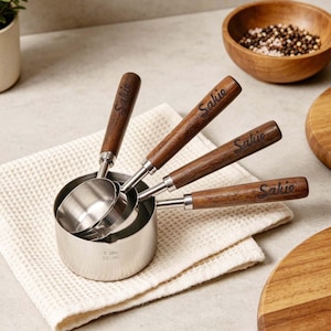 May include: A set of four stainless steel measuring cups with wooden handles. The handles are engraved with the word "Sakie". The cups are nested on a white waffle-weave cloth. A wooden bowl with peppercorns is in the background.