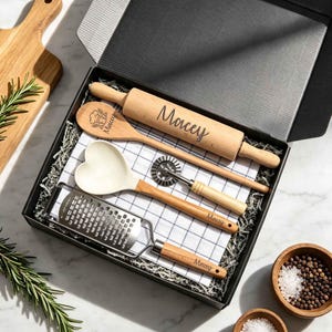 May include: A black gift box containing a wooden rolling pin, a wooden spoon, a heart-shaped silicone spatula, a metal pastry wheel, and a metal grater, all featuring the name Macey. A white and black grid patterned kitchen towel is included.
