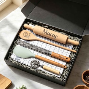 May include: A black gift box containing a wooden rolling pin, a wooden spoon, a metal grater, a sage green silicone spatula, and a pastry wheel, all personalized with the name Macey. A white and blue checkered kitchen towel is included.