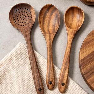K&ouml;nnte beinhalten: Drei K&uuml;chenutensilien aus Holz: ein Sch&ouml;pfl&ouml;ffel, ein normaler L&ouml;ffel und eine Sch&ouml;pfkelle. Die L&ouml;ffel sind aus dunkelbraunem Holz mit sichtbaren Maserungen gefertigt. Jeder L&ouml;ffel hat einen langen Griff und eine dunkle Niete am Ende des Griffs. Sie liegen auf einem wei&szlig;en Waffelstoff.