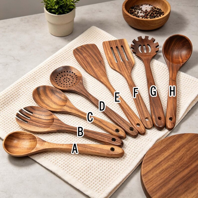 K&ouml;nnte beinhalten: Eine Sammlung von acht h&ouml;lzernen K&uuml;chenutensilien, darunter L&ouml;ffel, Spatel und ein Nudelsieb. Die Utensilien sind aus warm get&ouml;ntem Holz gefertigt und auf einem wei&szlig;en Tuch angeordnet. Jedes Utensil hat ein Loch im Griff zum Aufh&auml;ngen.