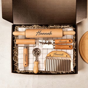 May include: A wooden rolling pin, pizza cutter, and other kitchen tools are arranged in a gift box. The rolling pin and other tools are engraved with the name "Hannah". The tools are on a checkered cloth.