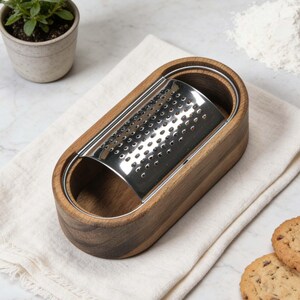 May include: A stainless steel grater with a wooden base, resting on a white cloth. The grater is oval-shaped and features a series of small holes for grating. A small potted plant and cookies are in the background. A kitchen utensil.