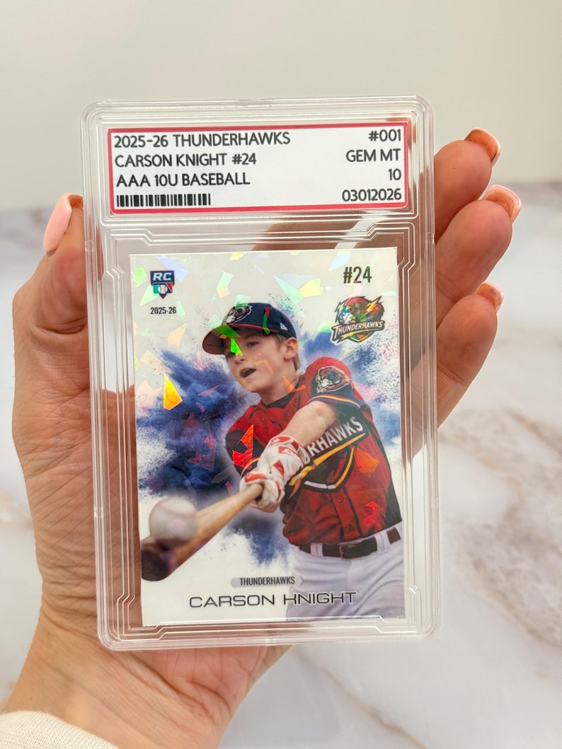 May include: A baseball trading card encased in a clear plastic holder. The card features a young baseball player in a red and black uniform swinging a bat. Text on the card includes "2025-26 Thunderhawks" and "Carson Knight #24".