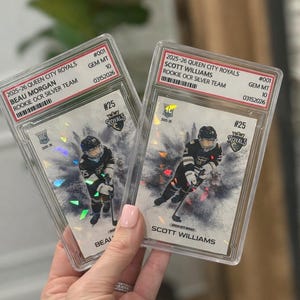 May include: Two graded hockey cards featuring players from the 2025-26 Queen City Royals. One card showcases Beau Morgan, the other Scott Williams. Both cards are graded GEM MT 10, with serial numbers. Each card has a player image.