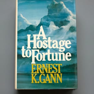Può includere: Un libro vintage con copertina rigida intitolato "A Hostage to Fortune" di Ernest K. Gann. La copertina presenta un paesaggio marino con nuvole bianche e acqua blu. Il titolo è in bianco e il nome dell'autore in giallo.