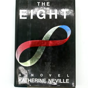 Puede incluir: Un libro de tapa dura negro titulado "The Eight" de Katherine Neville. El título está en blanco. Un símbolo de infinito colorido en rojo, azul y verde adorna el centro de la portada.