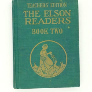 Puede incluir: Un libro vintage de color verde azulado titulado "The Elson Readers Book Two, Teachers' Edition". La portada presenta letras doradas y una ilustración circular de una niña alimentando gansos. El libro tiene un aspecto texturizado.