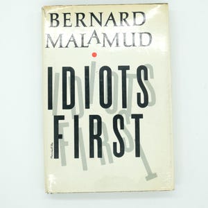 May include: A hardcover book titled "Idiots First" by Bernard Malamud. The title is in large, bold, black letters, with a shadow effect. The author's name is in a smaller, gray font. The book has a cream-colored cover with a red dot.