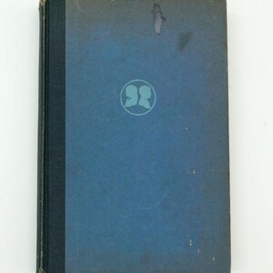 May include: A vintage book with a blue cover and a dark spine. The cover features a circular emblem with two silhouettes. The book's cover has some discoloration. The book is likely a collectible item.