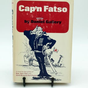 May include: A hardcover book titled Cap'n Fatso by Daniel Gallery with a white cover featuring a red rectangular block at the top. The illustration shows a saluting sailor in a dark uniform with a sword, a ship, and a small flag with a hammer and sickle.