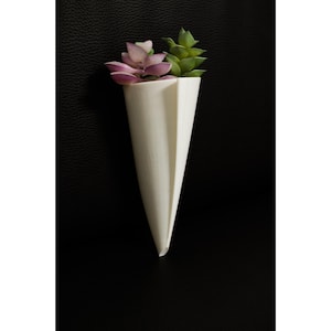 Könnte beinhalten: Ein weißer, kegelförmiger Wandtopf mit gefaltetem Design, der künstliche Sukkulenten enthält. Die Sukkulenten haben lila und grüne Blätter. Der Topf steht vor einem schwarzen Hintergrund.