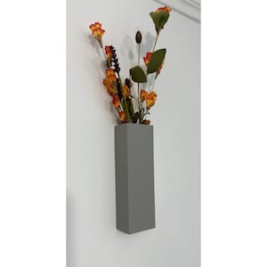 Puede incluir: Un jarrón de pared gris rectangular lleno de flores artificiales. Las flores son una mezcla de naranja, amarillo y burdeos, con hojas verdes. El jarrón está montado en una pared blanca, creando una exhibición floral moderna.