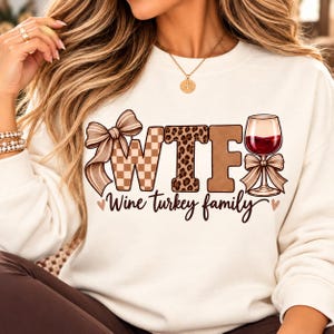 May include: A cream-colored sweatshirt featuring the text "WTF" in large letters with a tan checkered pattern, leopard print, and solid brown fill. A wine glass replaces the letter I, and the text "Wine turkey family" is printed below.