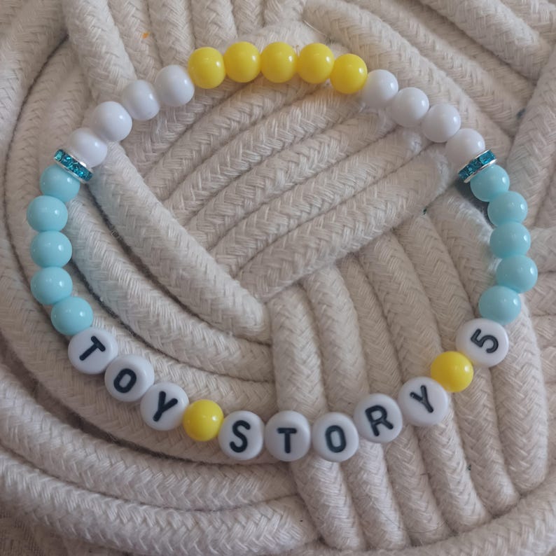 Puede incluir: Una pulsera circular de cuentas con esferas blancas, amarillas y azul claro. La pulsera incluye dos separadores plateados con cristales turquesa incrustados y cuentas con letras blancas que forman la frase TOY STORY 5.