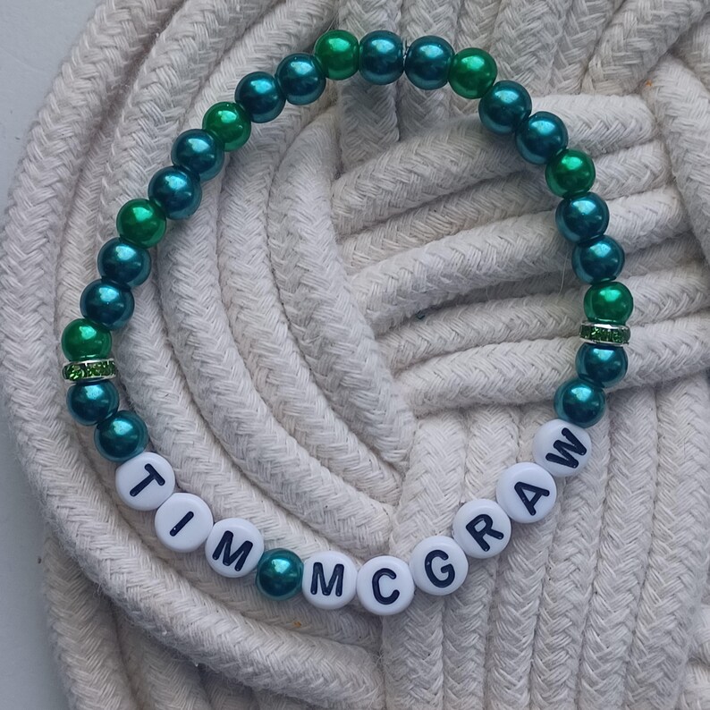 May include: A beaded bracelet with teal and green beads. The bracelet has white letter beads spelling out "TIM MCGRAW". The bracelet is on a white rope surface. The beads are a mix of teal and green, with a few clear rhinestone spacers.
