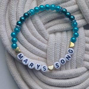May include: A beaded bracelet with turquoise beads and white letter beads spelling "MARYS SONG". The bracelet also features gold heart-shaped beads and silver rhinestone spacers. The bracelet is displayed on a white rope surface.