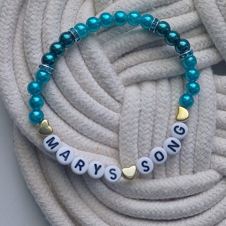 May include: A beaded bracelet with turquoise beads and white letter beads spelling "MARYS SONG". The bracelet also features gold heart-shaped beads and silver rhinestone spacers. The bracelet is displayed on a white rope surface.