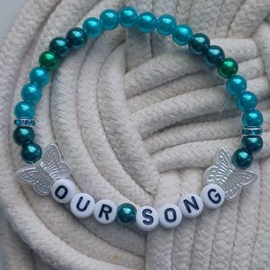 May include: A beaded bracelet with turquoise and dark green beads. The bracelet features the text "OUR SONG" spelled out with white letter beads. Two silver butterfly charms and rhinestone accents add detail.