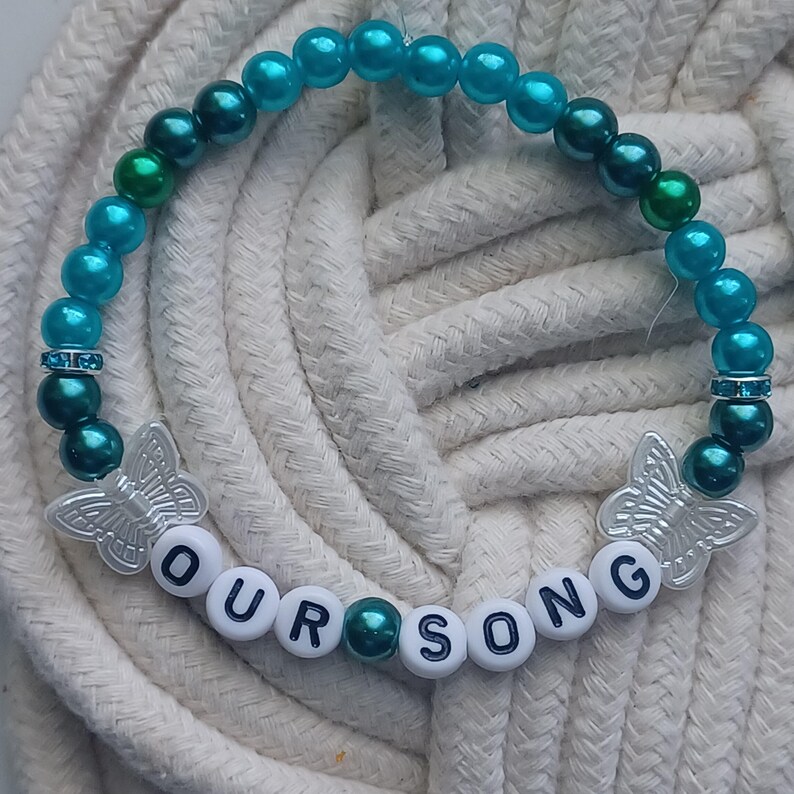 May include: A beaded bracelet with turquoise and dark green beads. The bracelet features the text "OUR SONG" spelled out with white letter beads. Two silver butterfly charms and rhinestone accents add detail.