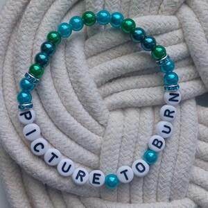 May include: A beaded bracelet with the words "PICTURE TO BURN" spelled out in white letter beads. The bracelet features teal, green, and blue beads, with rhinestone accents. The bracelet is resting on a white rope.