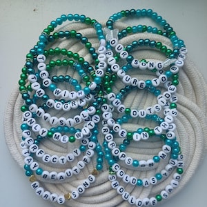 May include: A collection of beaded bracelets in shades of teal and green, with white letter beads spelling out song lyrics. The bracelets are arranged on a white, woven surface. Some bracelets have gold-colored heart charms.