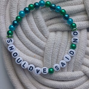 May include: A beaded bracelet with the words "SHOULD'VE SAID NO" spelled out in white letter beads. The bracelet features teal, green, and white beads, with green accents and a rhinestone spacer.
