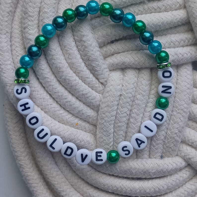 May include: A beaded bracelet with the words "SHOULD'VE SAID NO" spelled out in white letter beads. The bracelet features teal, green, and white beads, with green accents and a rhinestone spacer.