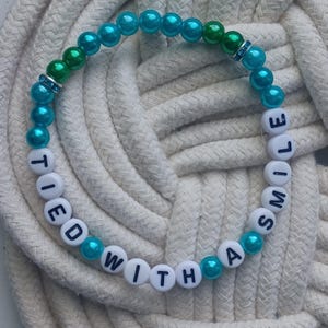 May include: A beaded bracelet with the words "TIED WITH A SMILE" spelled out in white letter beads. The bracelet features teal and green beads, with a few clear, sparkly accent beads. The bracelet is displayed on a white rope.