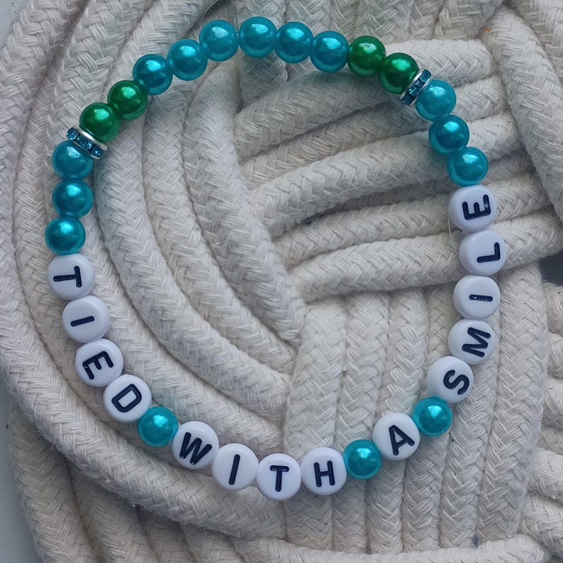 May include: A beaded bracelet with the words "TIED WITH A SMILE" spelled out in white letter beads. The bracelet features teal and green beads, with a few clear, sparkly accent beads. The bracelet is displayed on a white rope.