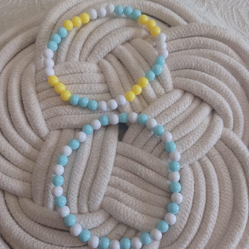 Puede incluir: Dos pulseras circulares de cuentas con un patr&oacute;n repetitivo de cuentas redondas y lisas en blanco, azul claro y amarillo. La pulsera superior alterna estos tres colores, mientras que la inferior presenta un patr&oacute;n alterno de cuentas blancas y azul claro.