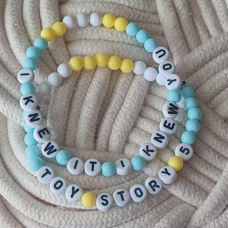Puede incluir: Dos pulseras circulares de cuentas con una mezcla de cuentas esf&eacute;ricas de color azul claro, blanco y amarillo. Una pulsera muestra el texto I KNEW IT en letras negras, mientras que la otra muestra el texto TOY STORY 5 en letras negras.