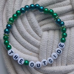 May include: A beaded bracelet with the words "THE OUTSIDE" spelled out in white letter beads. The bracelet features alternating green and teal beads, with rhinestone spacers.