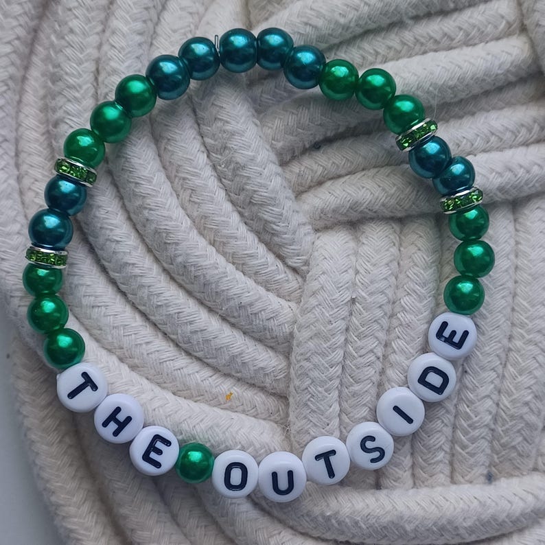 May include: A beaded bracelet with the words "THE OUTSIDE" spelled out in white letter beads. The bracelet features alternating green and teal beads, with rhinestone spacers.