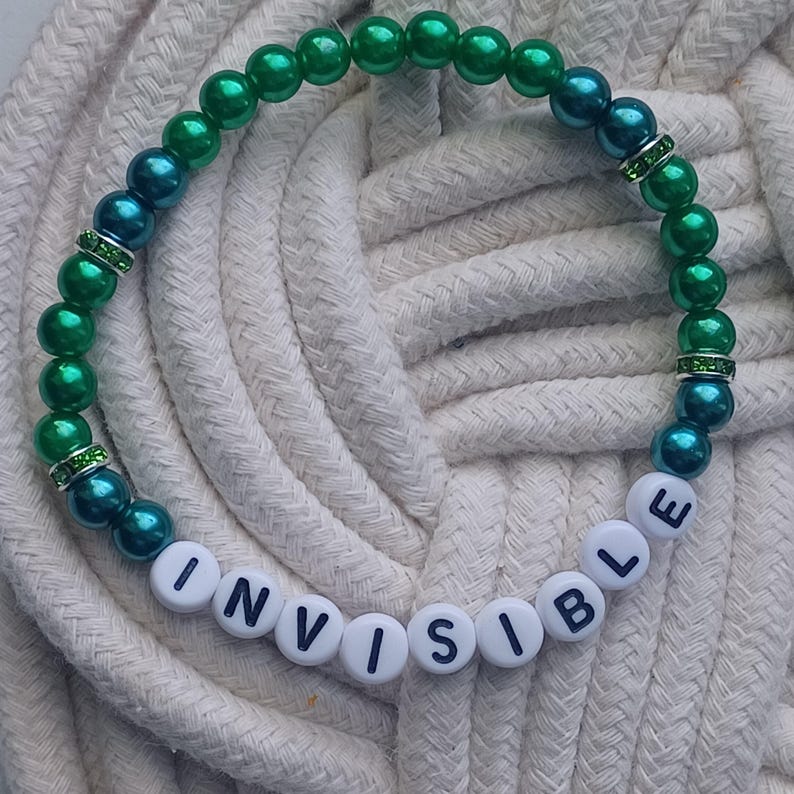 May include: A beaded bracelet with the word "INVISIBLE" spelled out in white letter beads. The bracelet features green and teal beads, with rhinestone accents. The bracelet is displayed on a white rope surface.