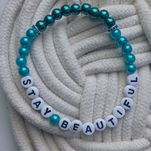 May include: A beaded bracelet with the words "STAY BEAUTIFUL" spelled out in white letter beads. The bracelet features teal and dark teal beads, with a small band of clear rhinestones. The bracelet is resting on a white rope surface.