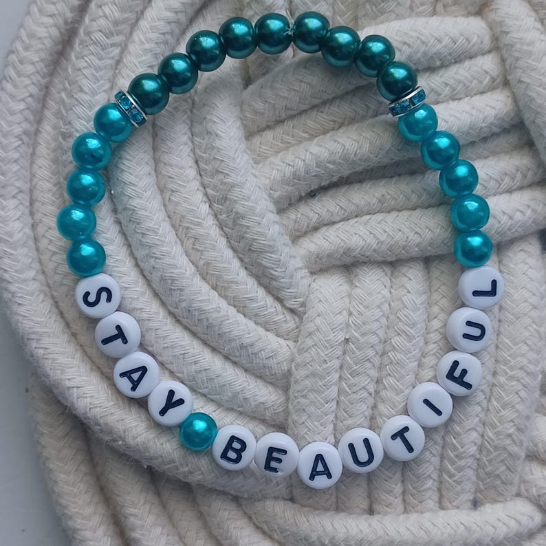 May include: A beaded bracelet with the words "STAY BEAUTIFUL" spelled out in white letter beads. The bracelet features teal and dark teal beads, with a small band of clear rhinestones. The bracelet is resting on a white rope surface.