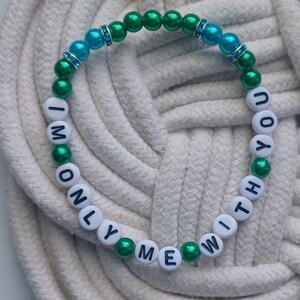 May include: A beaded bracelet with the words "I'M ONLY WITH YOU ME" spelled out in white letter beads. The bracelet features green and blue beads, with some blue beads accented with silver-colored spacers. The bracelet is on a white rope surface.