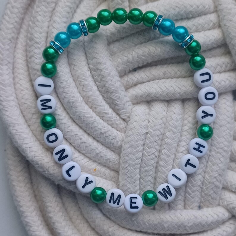 May include: A beaded bracelet with the words "I'M ONLY WITH YOU ME" spelled out in white letter beads. The bracelet features green and blue beads, with some blue beads accented with silver-colored spacers. The bracelet is on a white rope surface.