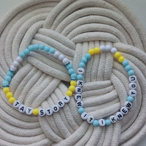 Puede incluir: Dos pulseras el&aacute;sticas de cuentas con abalorios redondos blancos, azul claro y amarillos. Una pulsera muestra el texto negro TAY STORY, mientras que la otra muestra el texto negro I KNEW IT I KNEW YOU. Ambas pulseras incluyen peque&ntilde;os detalles azules.